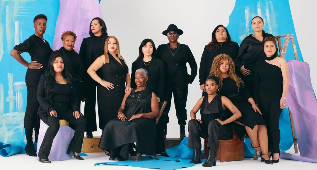 Group of diverse women wearing black clothing posing together against a blue and purple backdrop.