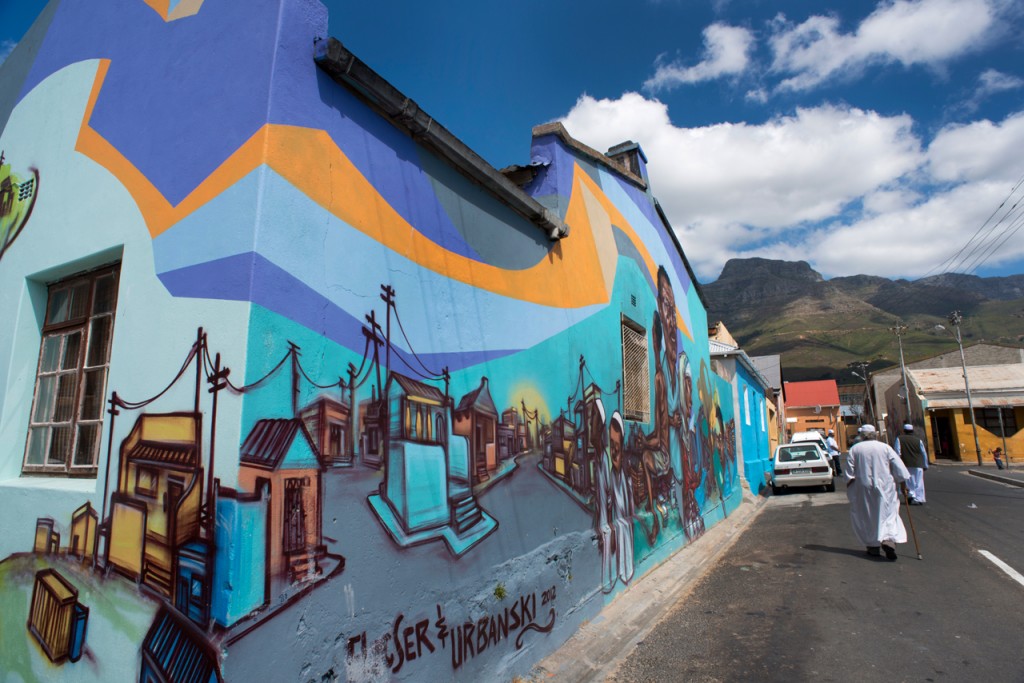 Street art mural on a blue building in Woodstock, Cape Town, depicting stylized houses and buildings with Table Mountain visible in the background and a person walking on the street. 