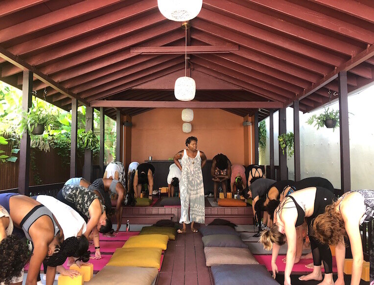 A diverse group of people practicing yoga at HealHaus in Brooklyn, guided by a Black female instructor under a covered outdoor pavilion.