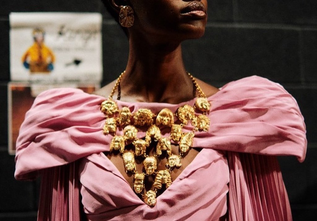 Model wearing pink draped dress with gold face-pendant necklace from Johnny Nelson Jewelry.Caption: 