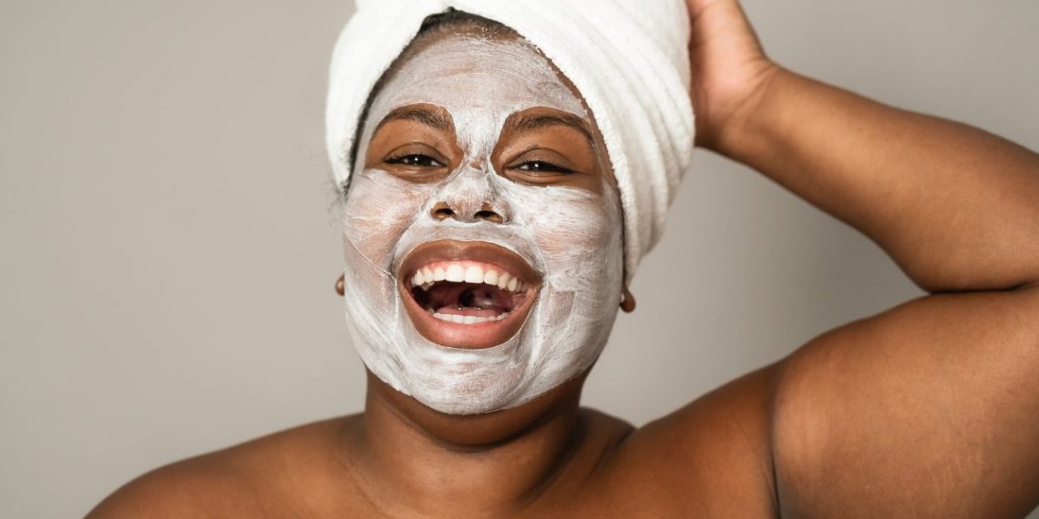 Black woman smiling joyfully with a towel on her head and a clay face mask, embracing self-care.