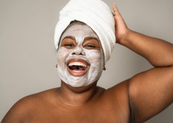 Black woman smiling joyfully with a towel on her head and a clay face mask, embracing self-care.