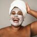 Black woman smiling joyfully with a towel on her head and a clay face mask, embracing self-care.