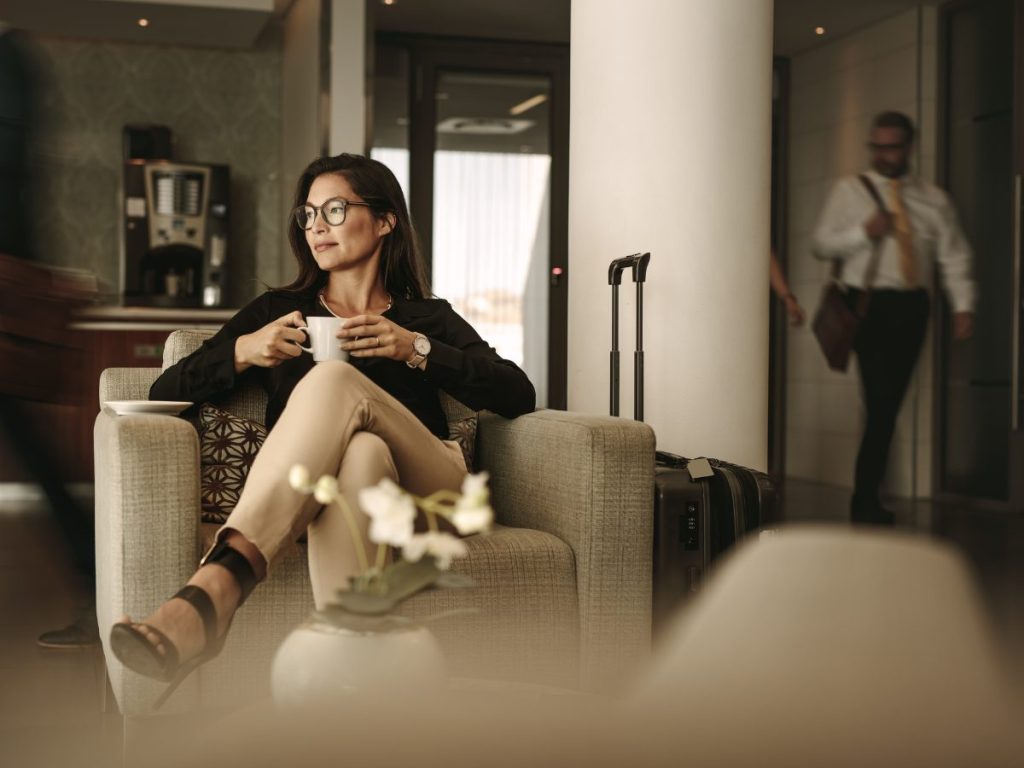 A woman sitting comfortably in an airport lounge, looking relaxed and focused on travel recovery. She gazes out the window, enjoying a moment of calm before her flight, with a suitcase nearby.