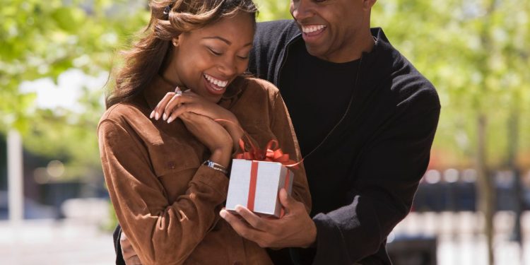 Black couple smiling as he gives her a gift, showcasing.