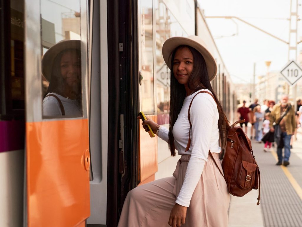 A solo Black woman wearing a beige hat and white top steps onto a train, holding a yellow ticket in her hand while carrying a brown backpack, with a busy platform in the background.