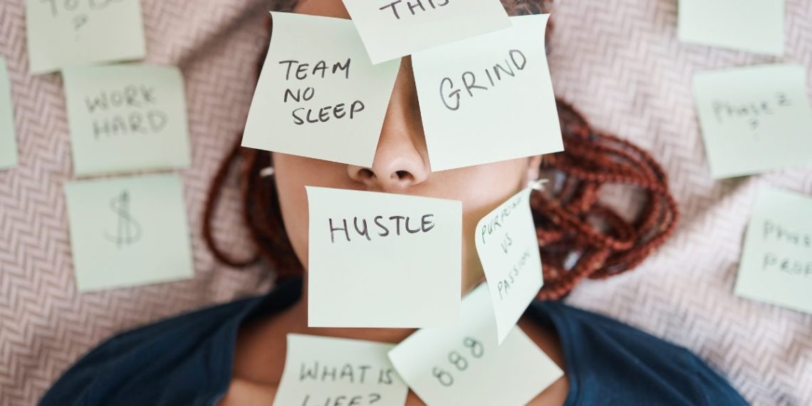 Overwhelmed woman lying on a bed with sticky notes that say "Hustle," "Grind," and "No Sleep," representing burnout from hustle culture.