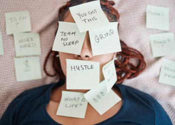 Overwhelmed woman lying on a bed with sticky notes that say "Hustle," "Grind," and "No Sleep," representing burnout from hustle culture.