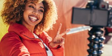 A smiling solo Black woman with curly hair takes a selfie in a red jacket, making a peace sign while standing in front of a colorful wall.