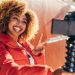 A smiling solo Black woman with curly hair takes a selfie in a red jacket, making a peace sign while standing in front of a colorful wall.