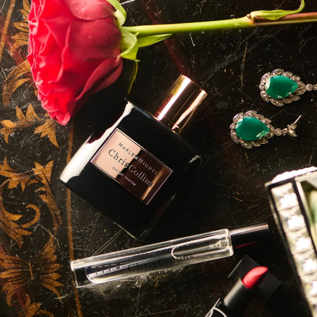 World of Chris Collins luxury perfume bottle (Harlem Nights) on black marble with gold accents, alongside a red rose, green gemstone earrings, lipstick, and perfume applicator.