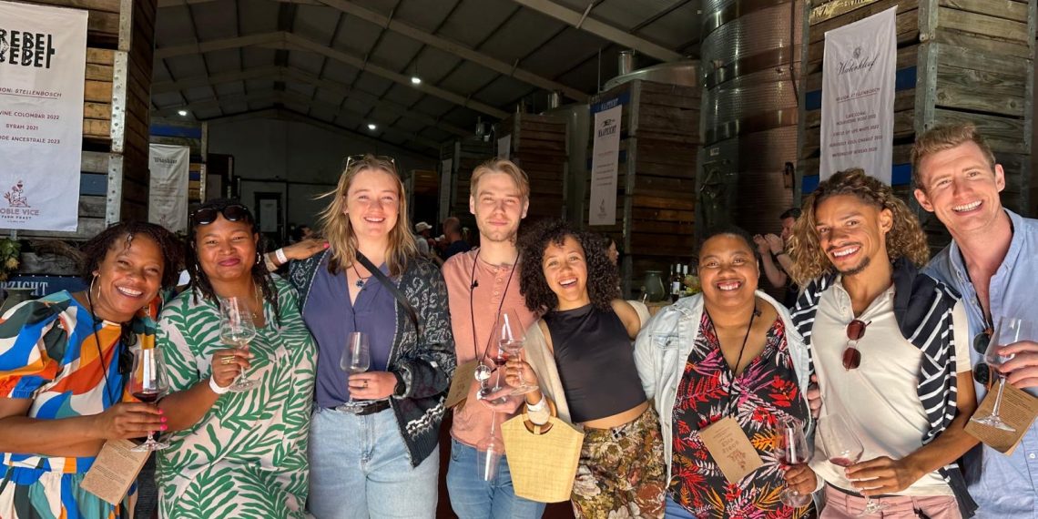 Wine festival lovers holding glasses and smiling together at an indoor tasting event with wine tanks and banners visible.
