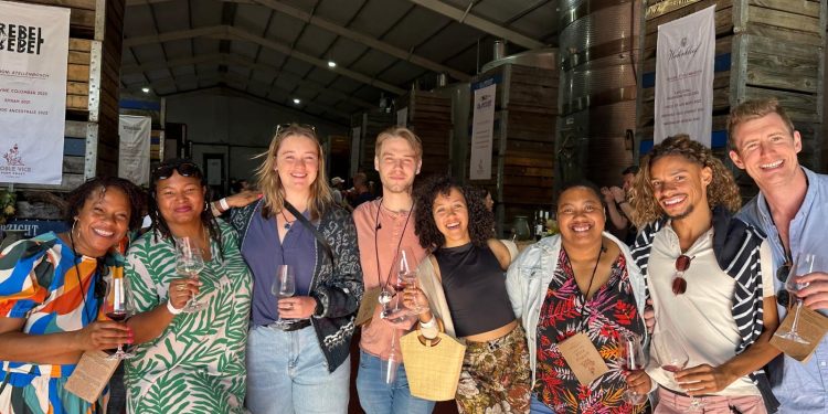 Wine festival lovers holding glasses and smiling together at an indoor tasting event with wine tanks and banners visible.