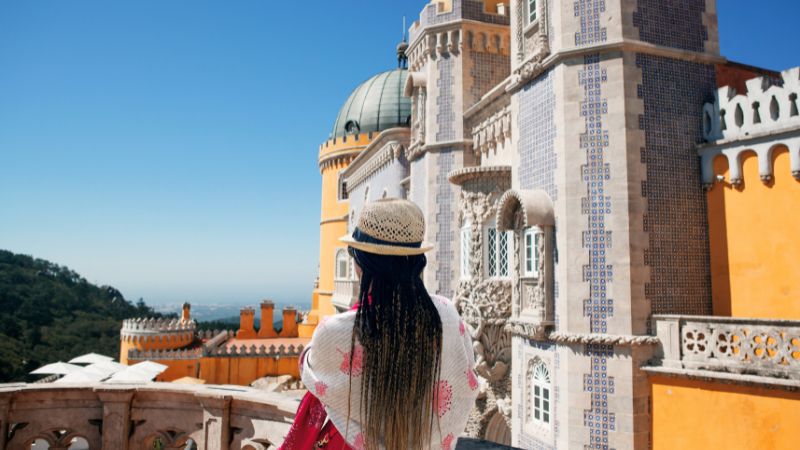 A woman wearing a straw hat and long braids gazes at the ornate, colorful facades and rooftop domes of Pena Palace in Sintra, Portugal, captured during her guided Douro River wine cruise adventure.