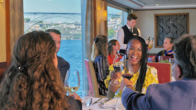 Guests on the AmaSintra cruise ship clink wine glasses during dinner, with the Douro River and a scenic bridge visible through large windows in the background.