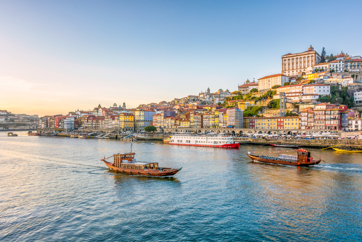 Scenic view of Porto, Portugal at sunset showing colorful historic buildings cascading down hillside to Douro River with traditional wooden boats in foreground. 