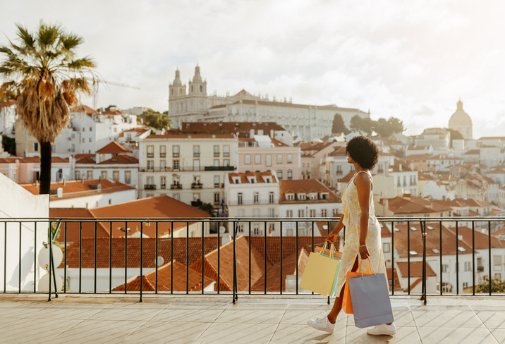 Woman in white dress holding luxury shopping bags on scenic terrace overlooking historic Lisbon cityscape with traditional red-tiled roofs, palm trees, and monastery in distance.