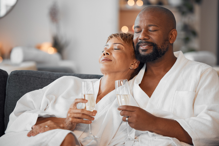Relaxed Black couple in white bathrobes holding champagne glasses with eyes closed, enjoying peaceful moment together on couch in luxury spa setting. 