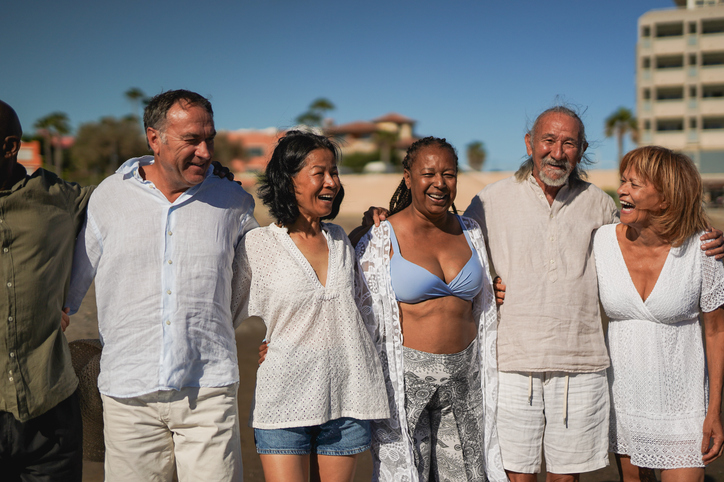 Group of diverse senior friends laughing and embracing on sunny beach, wearing casual white summer clothing with palm trees and resort buildings in background