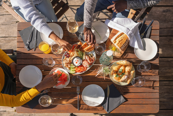 Overhead view of friends sharing gourmet lunch at wooden outdoor table with charcuterie boards, artisan bread, wine glasses and elegant place settings in sunny dining setting. 