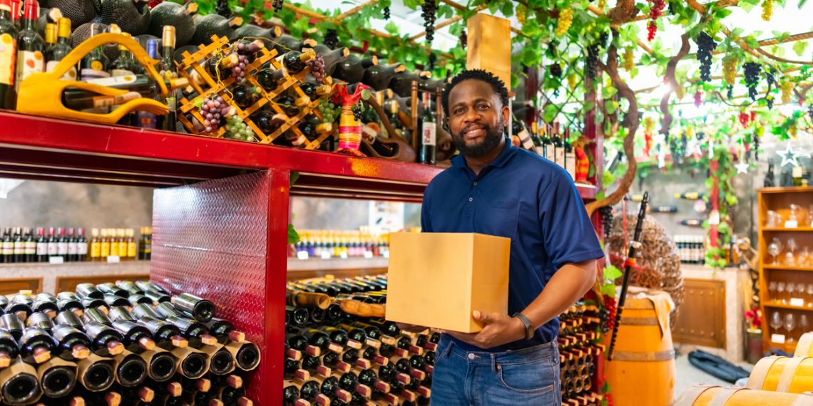 A smiling man standing inside a wine cellar surrounded by bottles and barrels, representing the growth and success of Black-owned wineries.