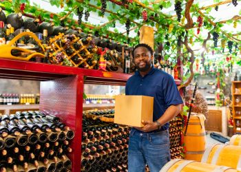 A smiling man standing inside a wine cellar surrounded by bottles and barrels, representing the growth and success of Black-owned wineries.