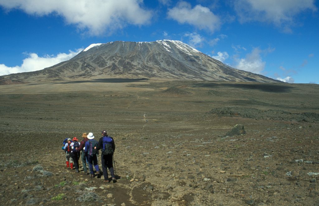 Group of hikers trekking toward Mount Kilimanjaro, Tanzania, with its snow-capped summit under blue skies.