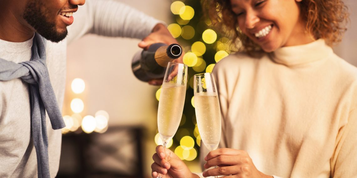 A smiling Black couple pours and shares glasses of sparkling wine during a festive celebration.