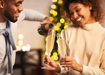 A smiling Black couple pours and shares glasses of sparkling wine during a festive celebration.