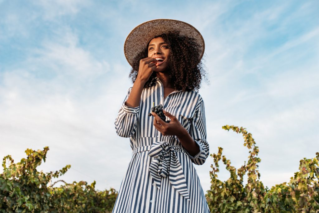 A smiling Black woman tastes grapes in a vineyard under a blue sky.